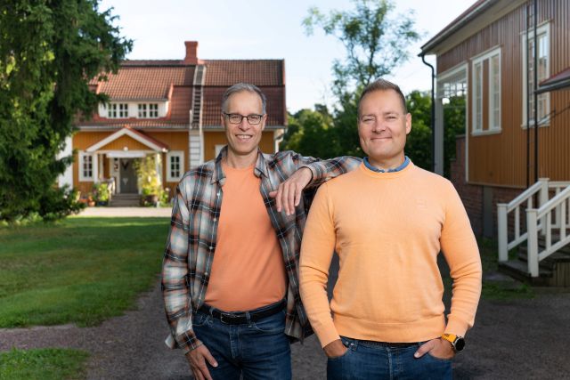 kaksi miestä seisoo vierekkäin puutalojen edessä