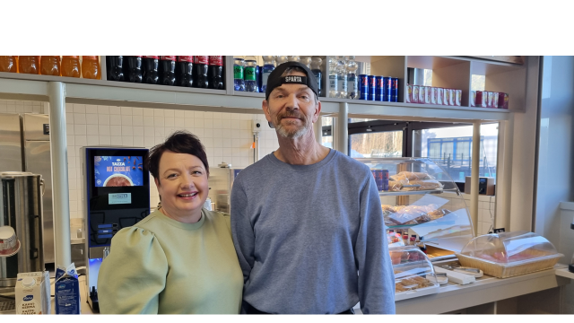 nainen ja mies seisomassa vierekkäin kahvilan tiskin edessä katsoen kameraan ja hymyillen