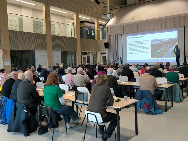 Ihmisiä koulun ruokasalissa pöytien ääressä seuraamassa taululle heijastettua esitystä