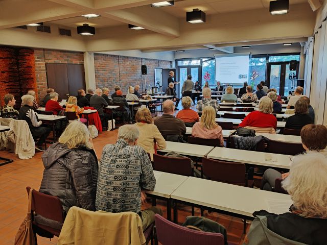 Yleisöä istumassa pöytien ääressä suuressa salissa ja kuuntelemassa esitystä terveysasemien kohtalosta