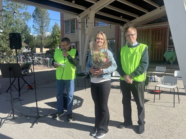 Kolme ihmistä seisomassa katoksen alla aurinkoisessa säässä, keskellä nainen, jolla kukkakimppu kädessä. Kuva Anitta Partanen